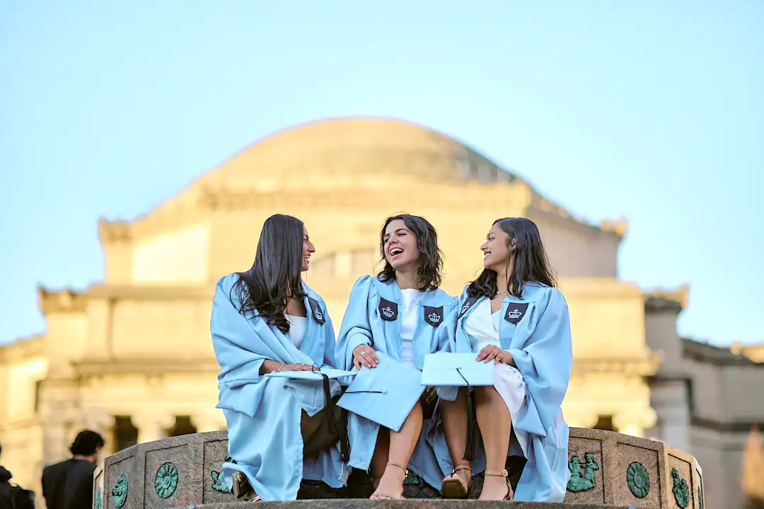 Graduate in white at golden hour leaning on railing, NYC skyline behind