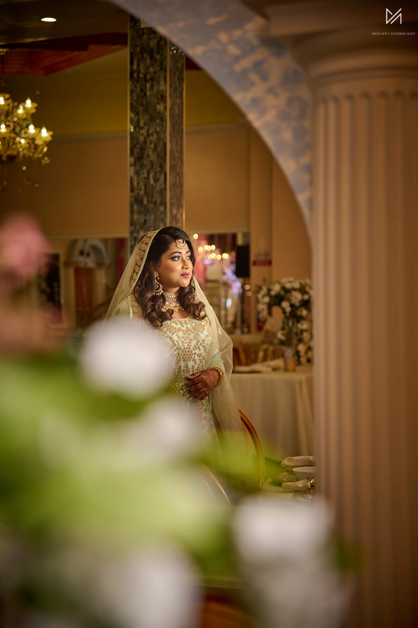 Bride in mint green and gold outfit framed by white roses in soft foreground focus