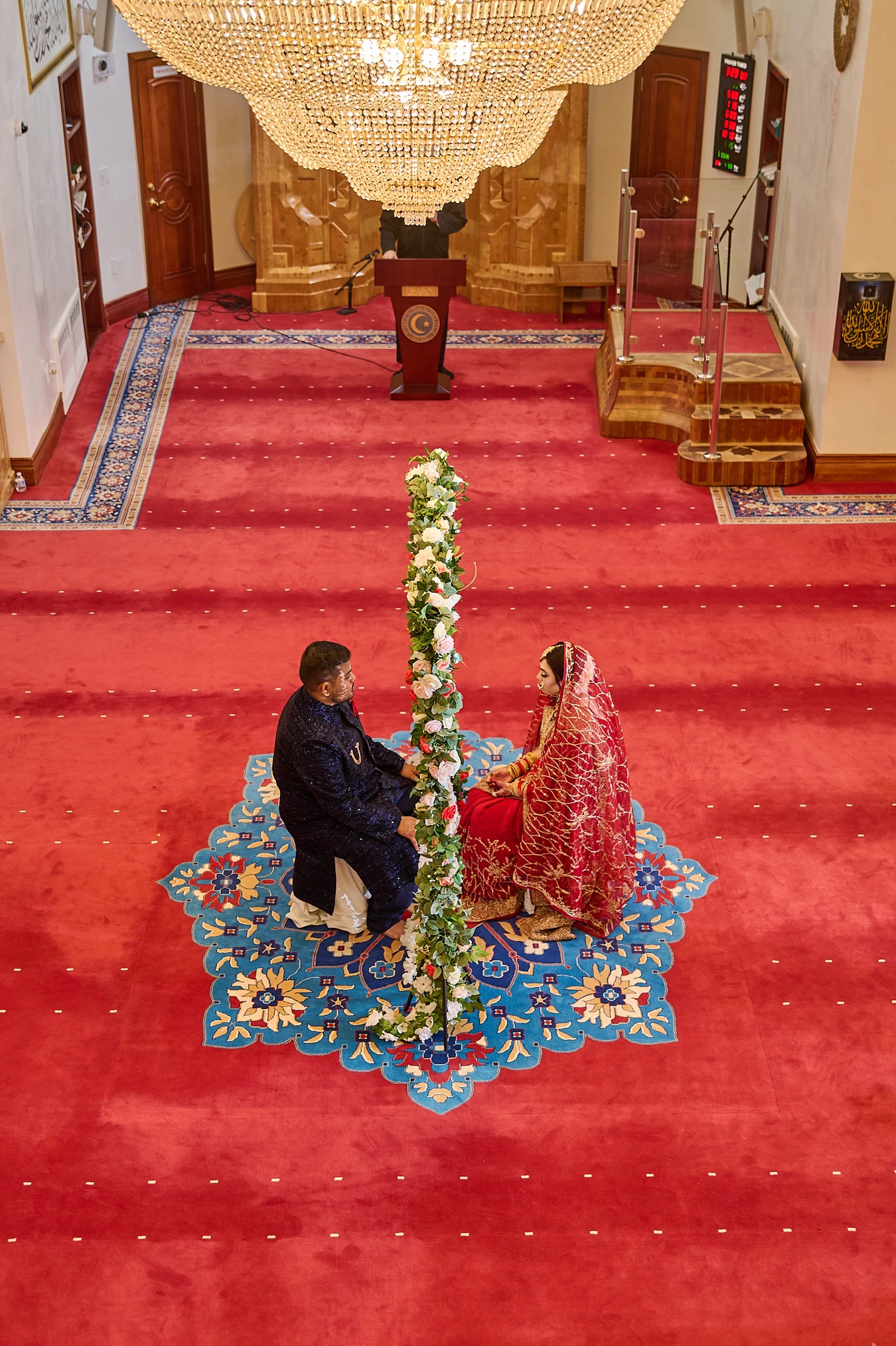 Aerial view of the Nikkah ceremony — bride in red and groom in navy on a patterned rug, separated by a floral garland, beneath a chandelier