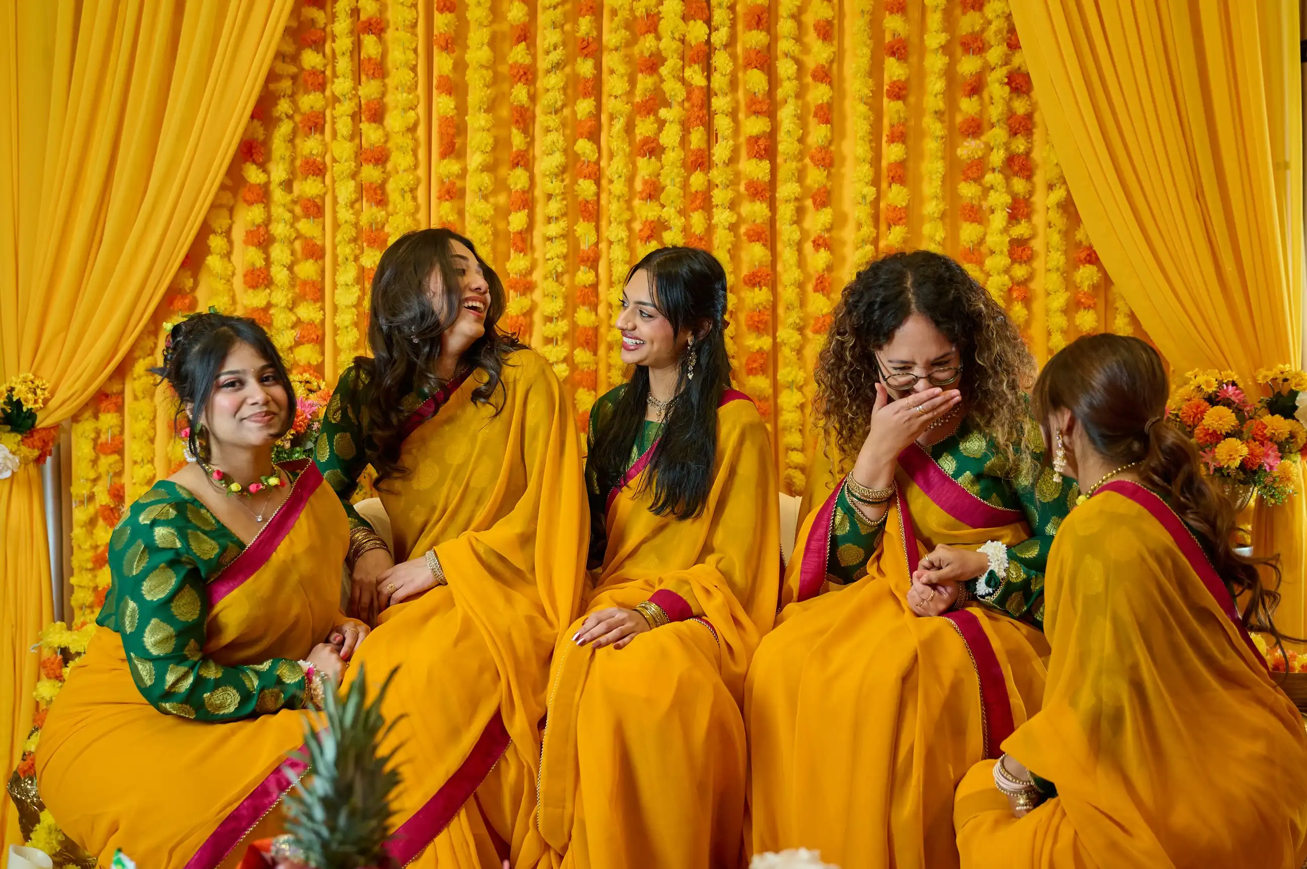 Five bridesmaids in yellow saris with green and red blouses laughing together against a marigold backdrop