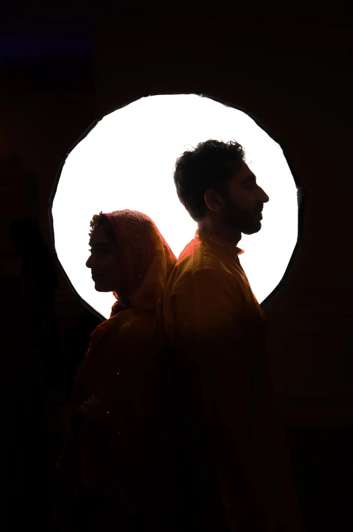 Silhouetted profiles of bride and groom back-to-back in front of an enormous round backlit disc